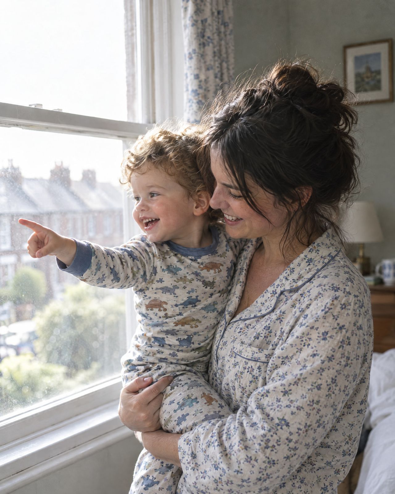 The mother holds her toddler by the window as he points outside.