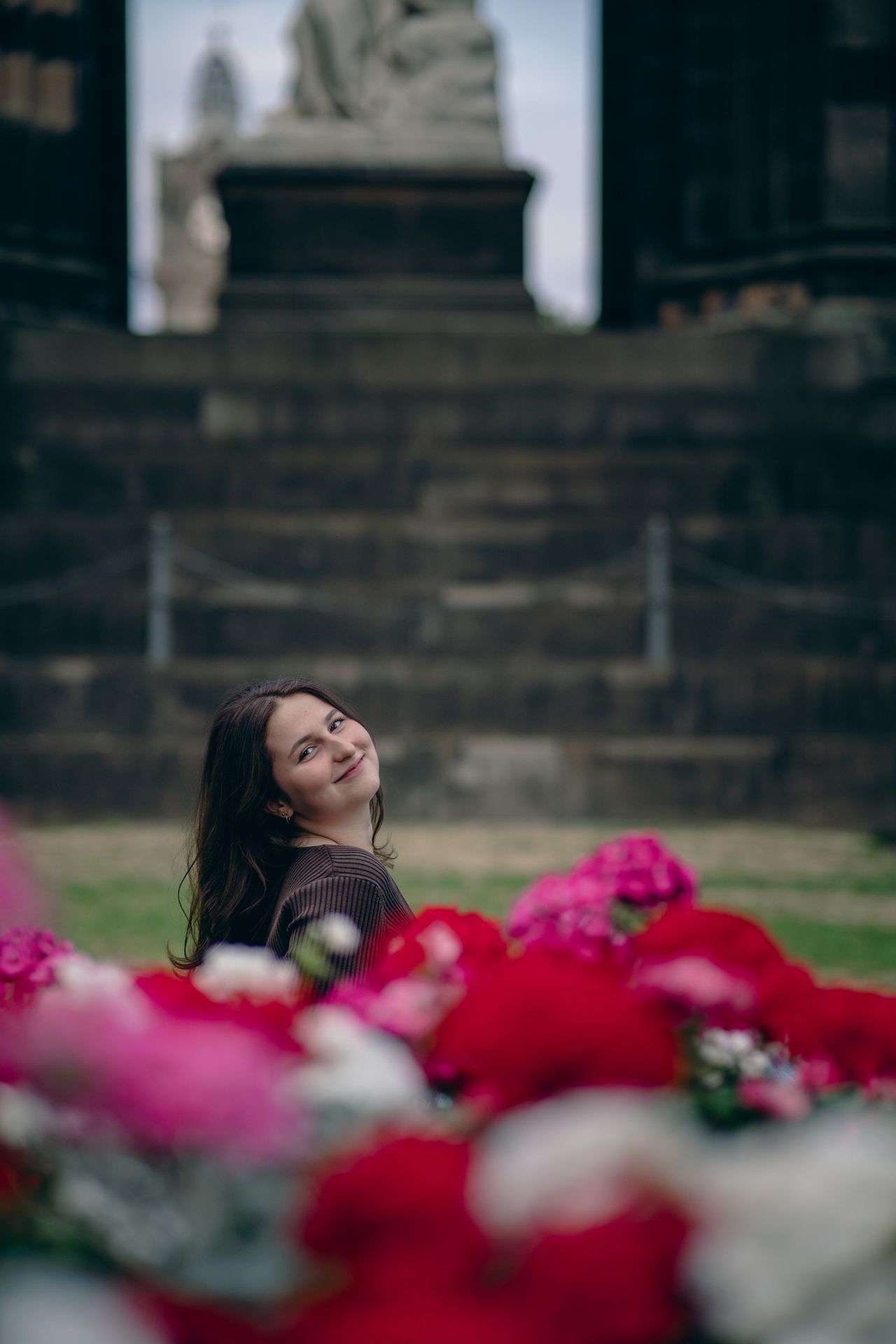 Smiling behind a bed of red and pink flowers, the steps of a monument behind.