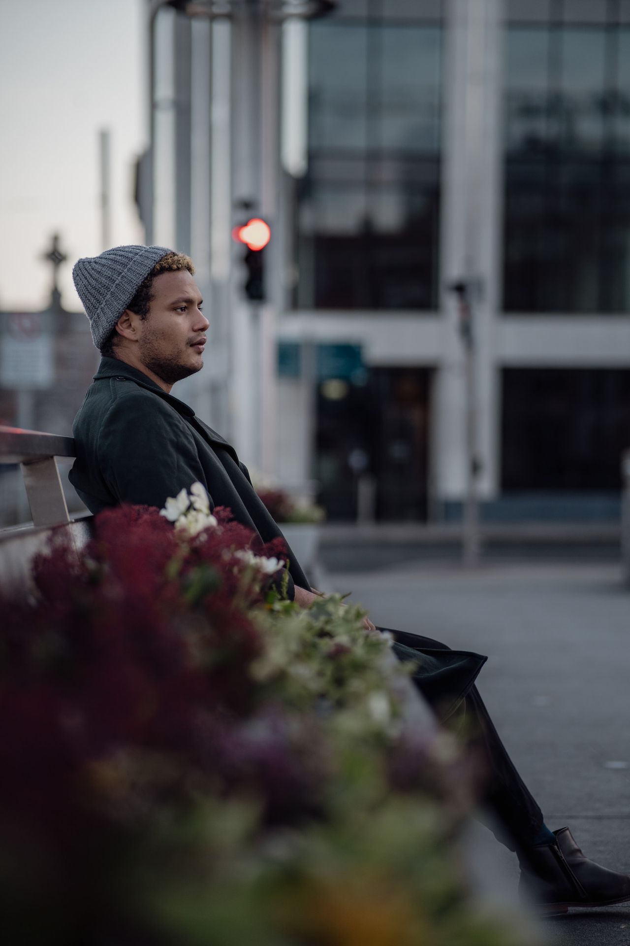 Sitting on a bench behind a planter of red flowers, looking into the distance.