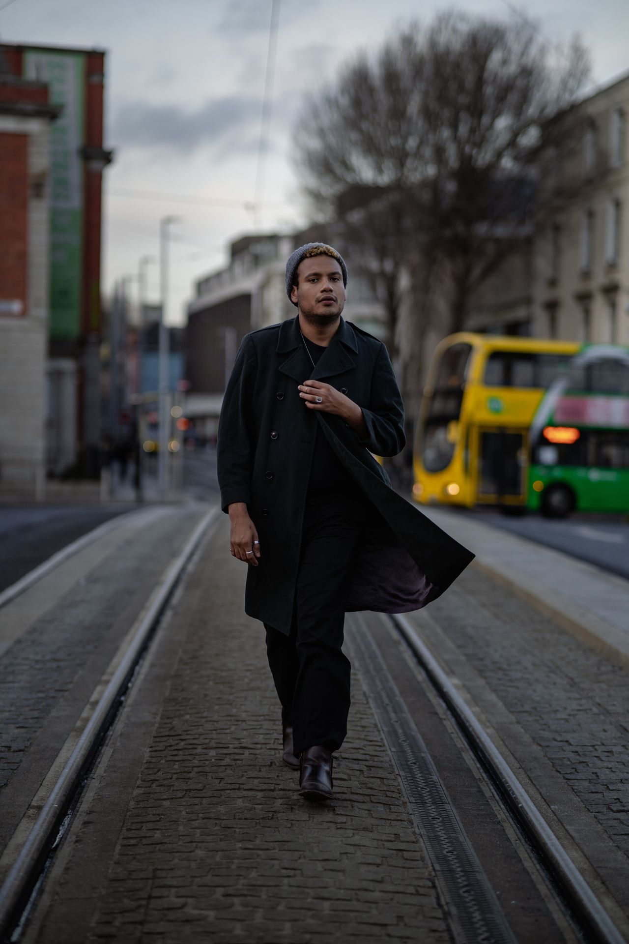 Walking along the tram tracks in a long dark coat, double-decker buses behind.