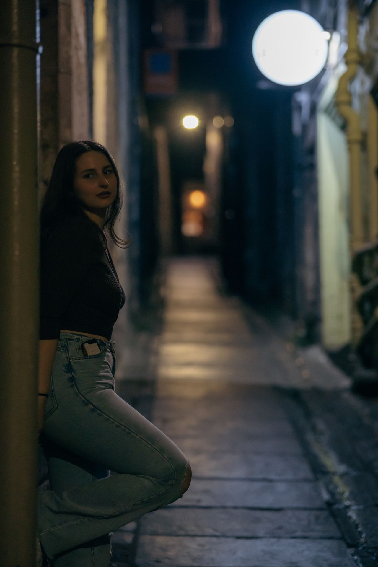 Leaning against the wall of a narrow lane at night, a round lamp glowing above.