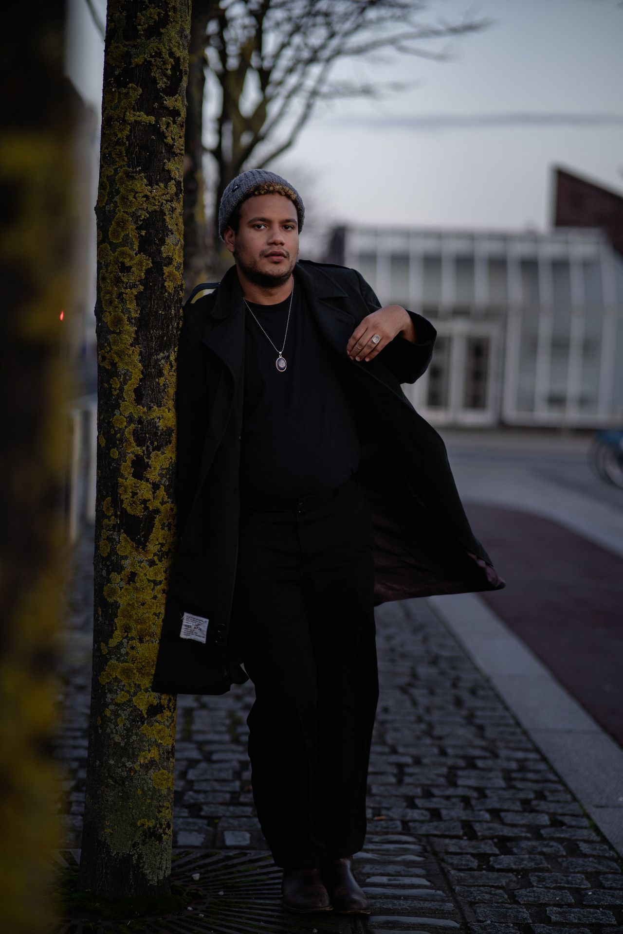 Leaning against a tree on a cobbled street, coat open, looking to one side.