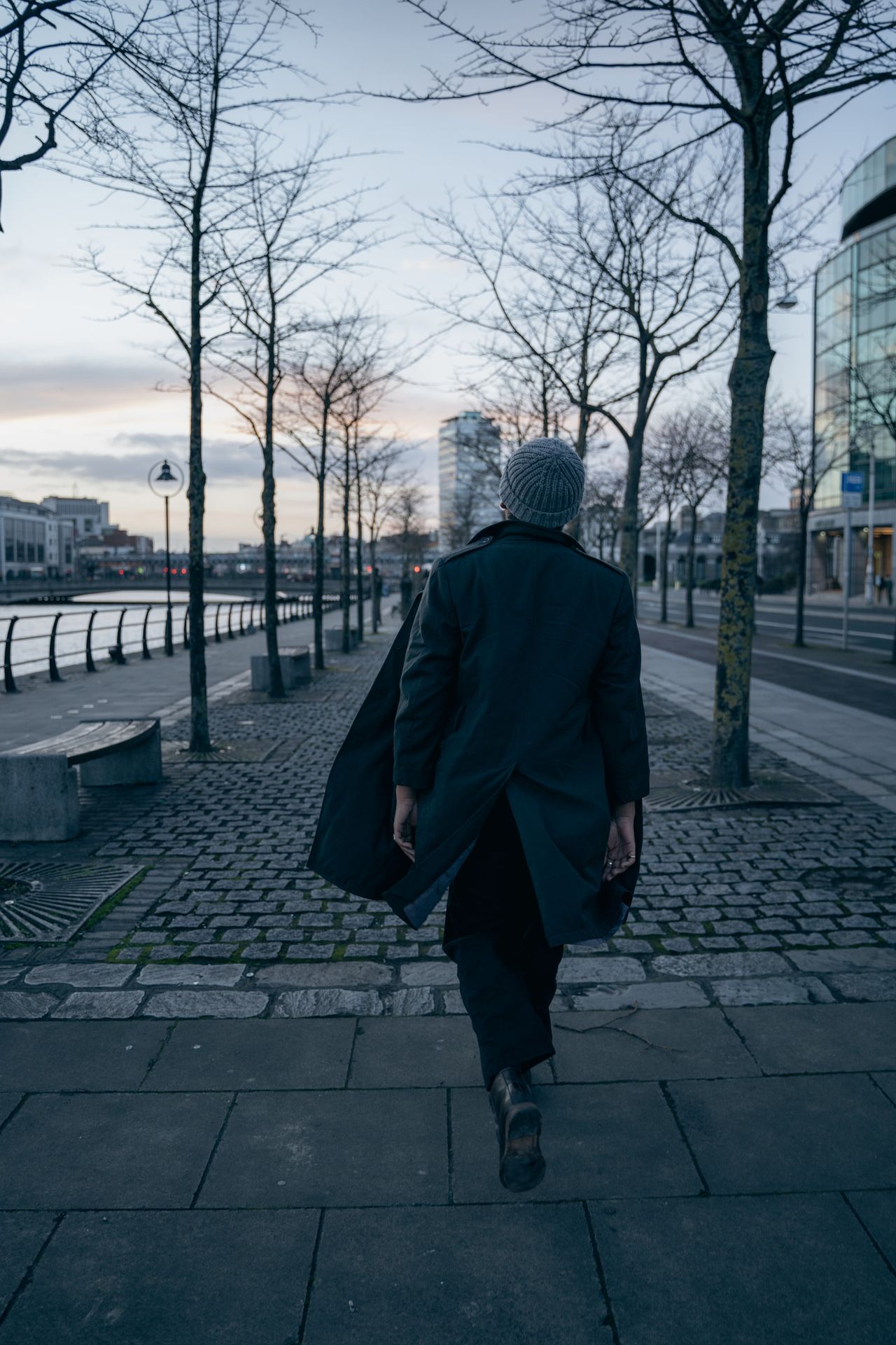 Walking away along a riverside promenade lined with bare trees.