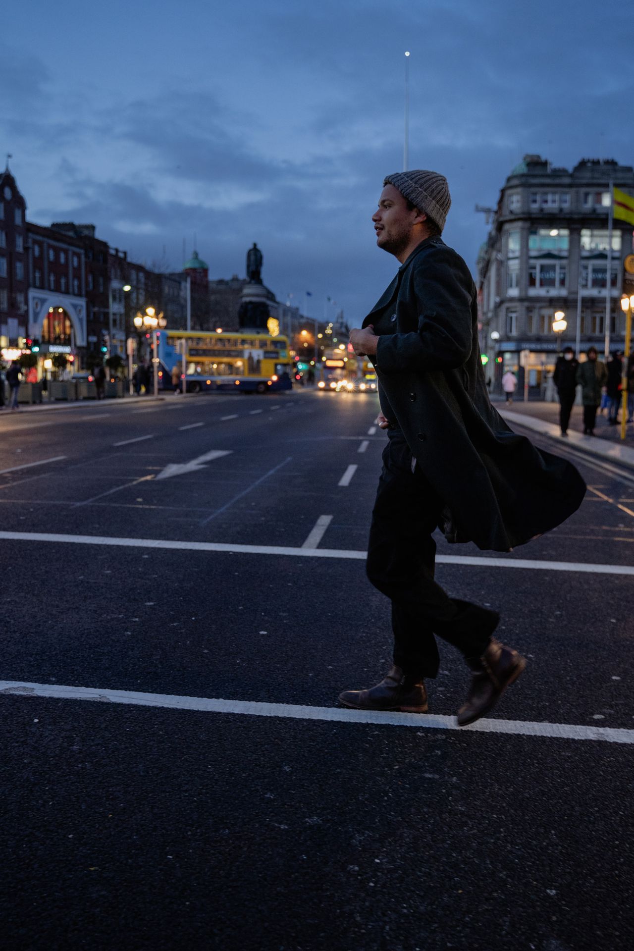 Crossing a wide street at blue hour, coat flying, the city lights coming on.