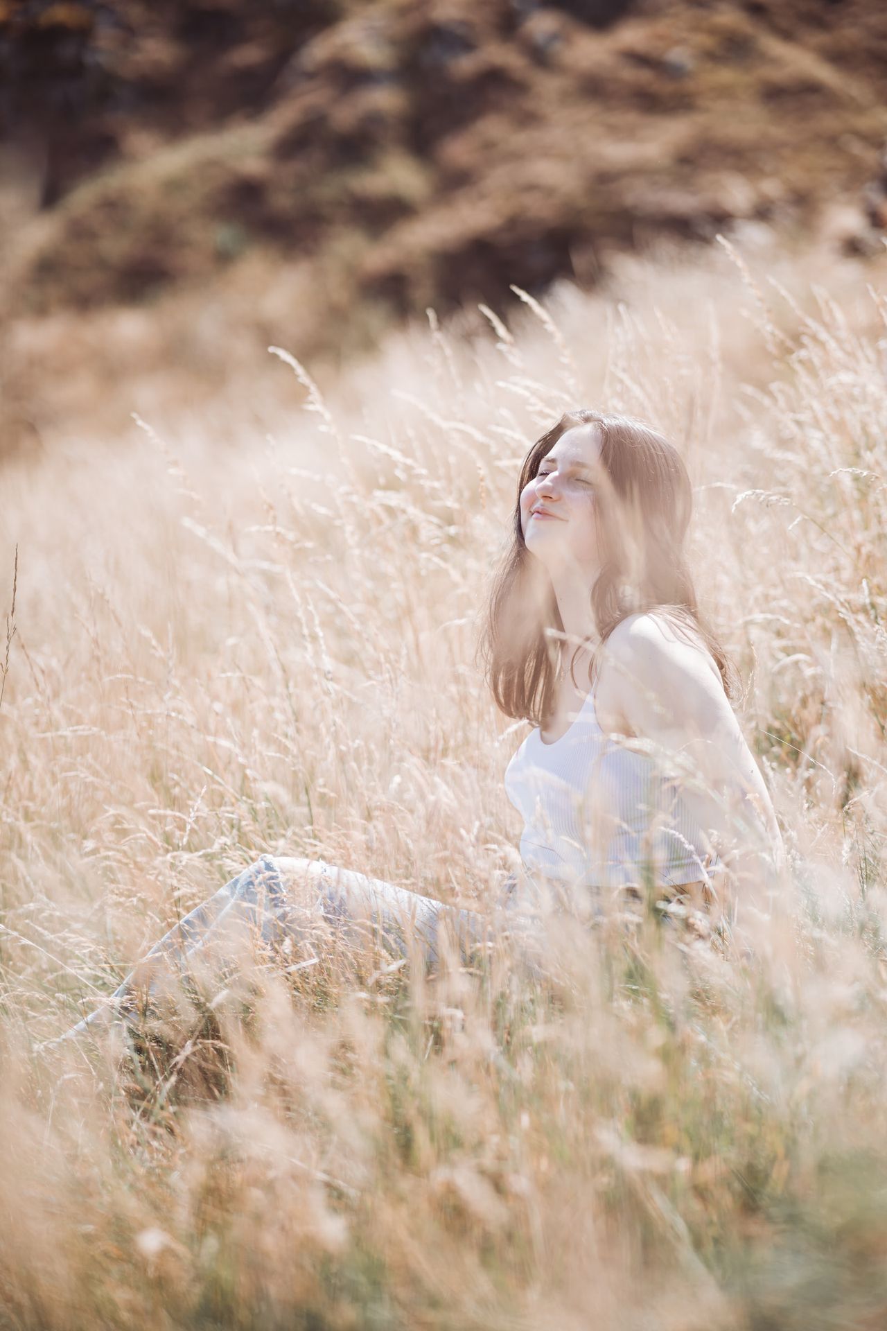 Sitting in tall golden grass in bright sun, eyes closed and smiling, a rocky hillside behind.