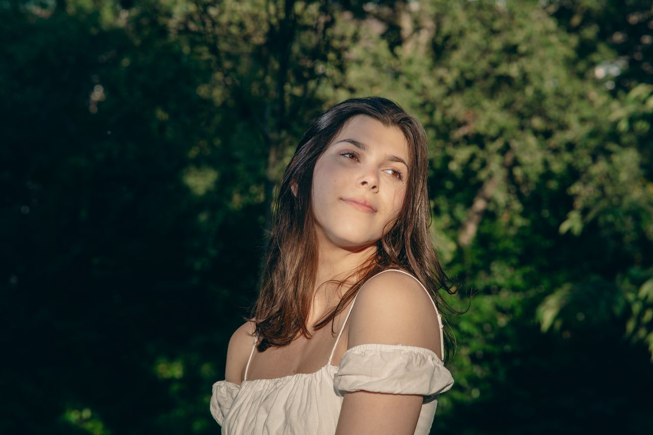 Looking over one shoulder in front of deep green trees, evening light on the face.