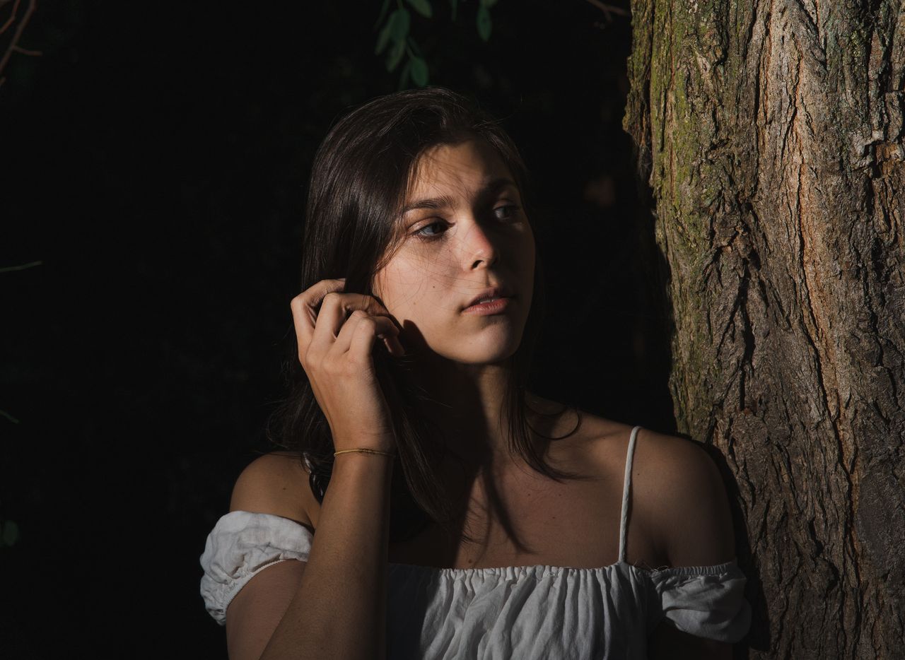 Leaning on a tree trunk in the shade, tucking the hair behind one ear.