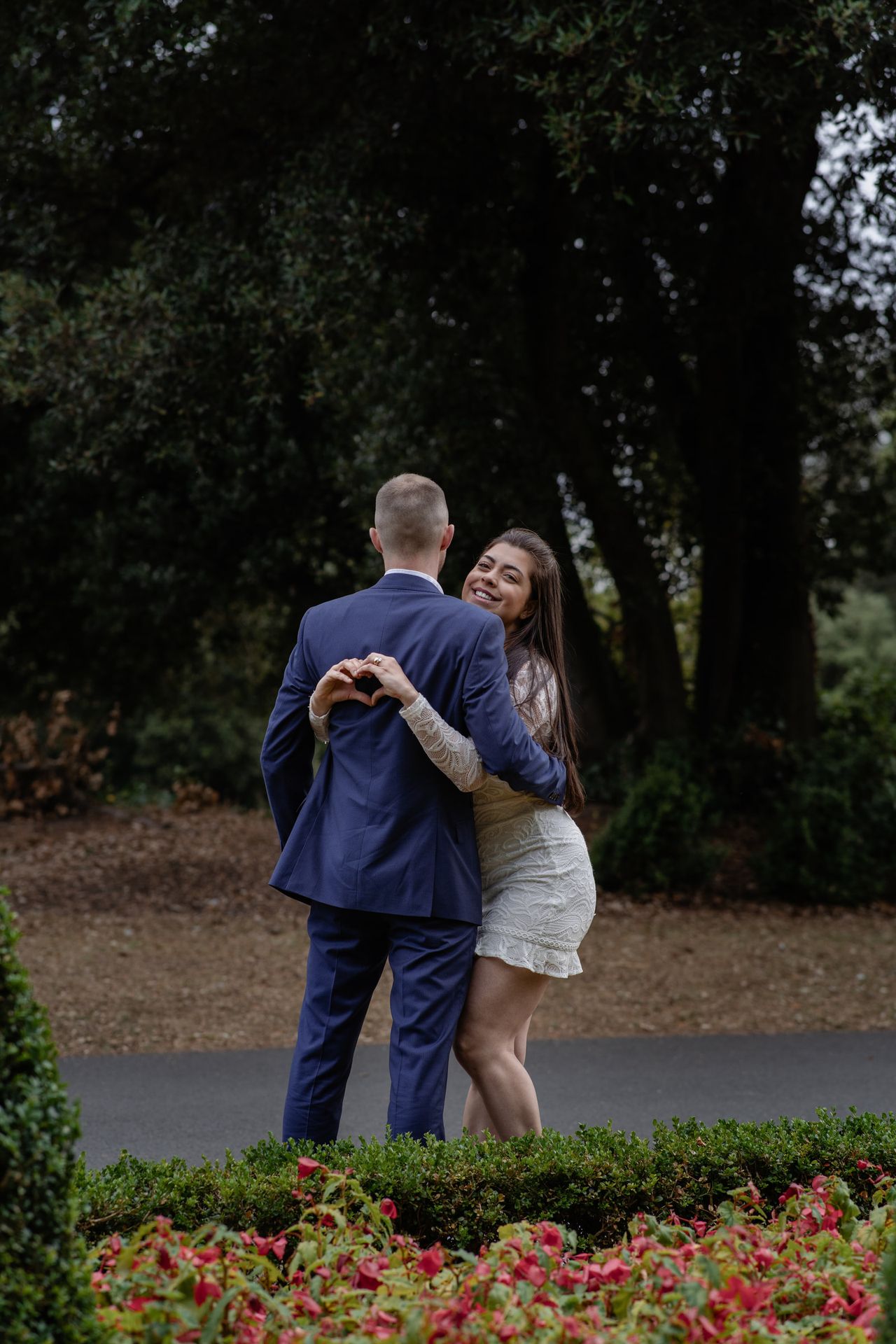 Arm in arm on a garden path under tall dark trees, the one in the white lace dress smiling back at the camera.