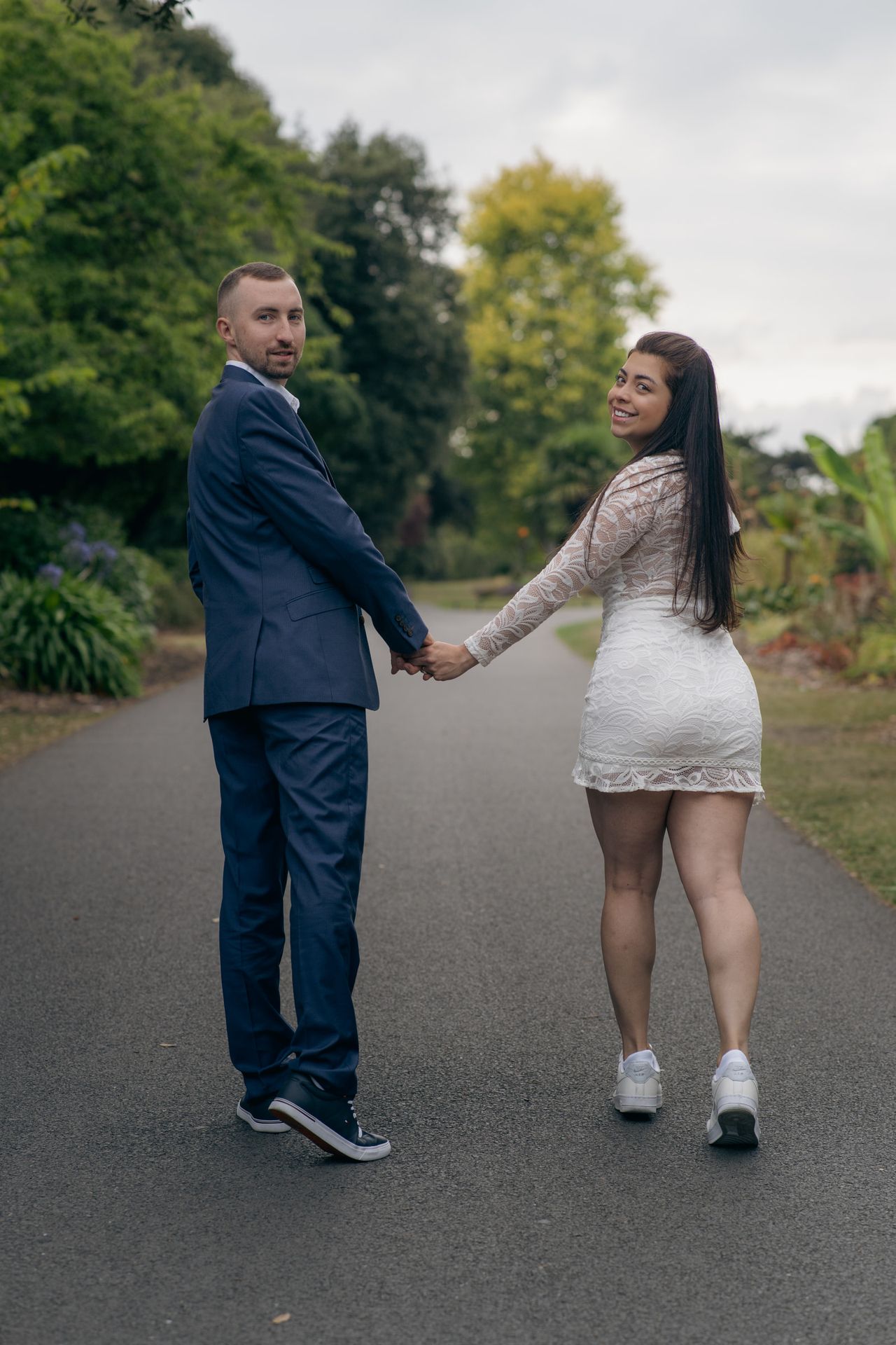 Holding hands on a park path, both looking back at the camera, in a navy suit and a white lace dress.