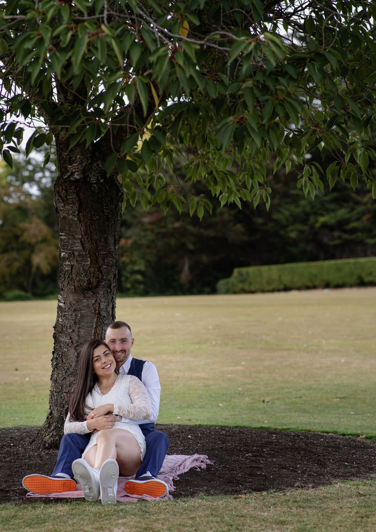 Sitting on a blanket at the foot of a tree in an embrace, an open lawn and hedges behind.