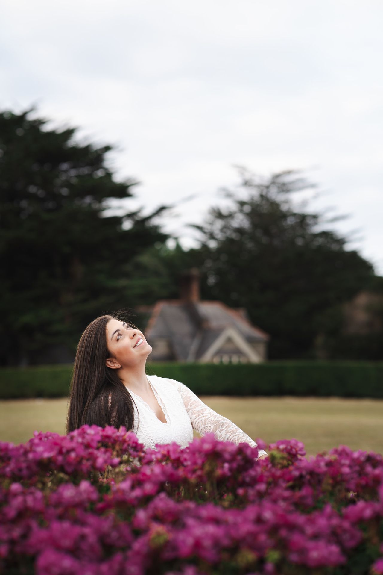 Laughing up at the sky behind a bank of magenta flowers, a gabled house among dark trees behind.