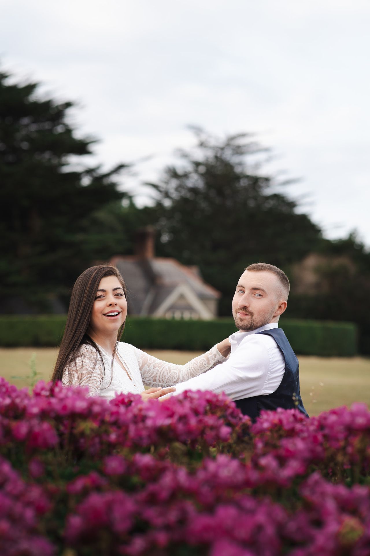 Laughing together behind the magenta flowers, one hand on the other’s shoulder.