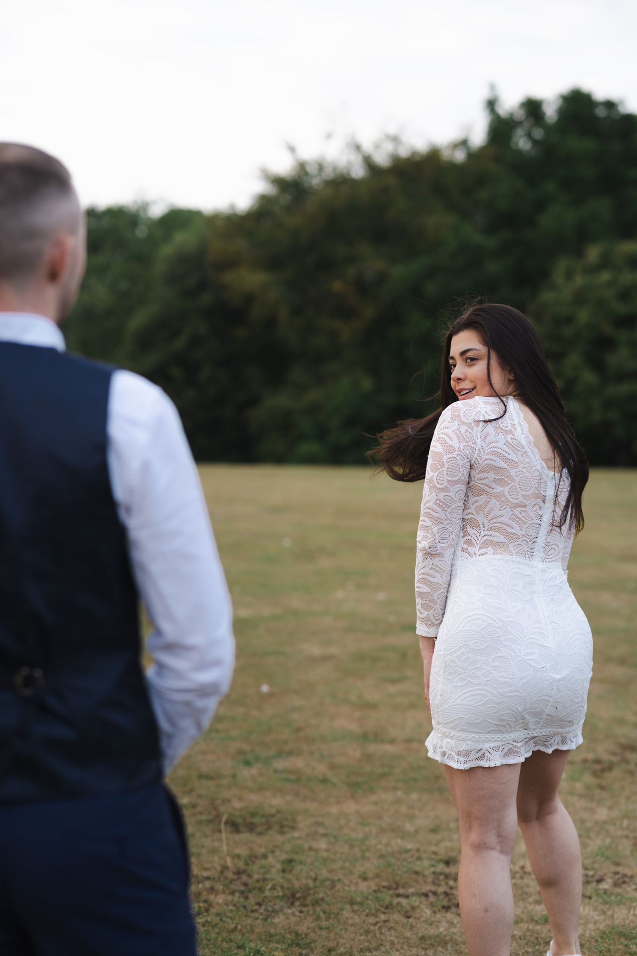 Past a navy waistcoat in the foreground, a turn back across the lawn in white lace, the hair lifted by the breeze.