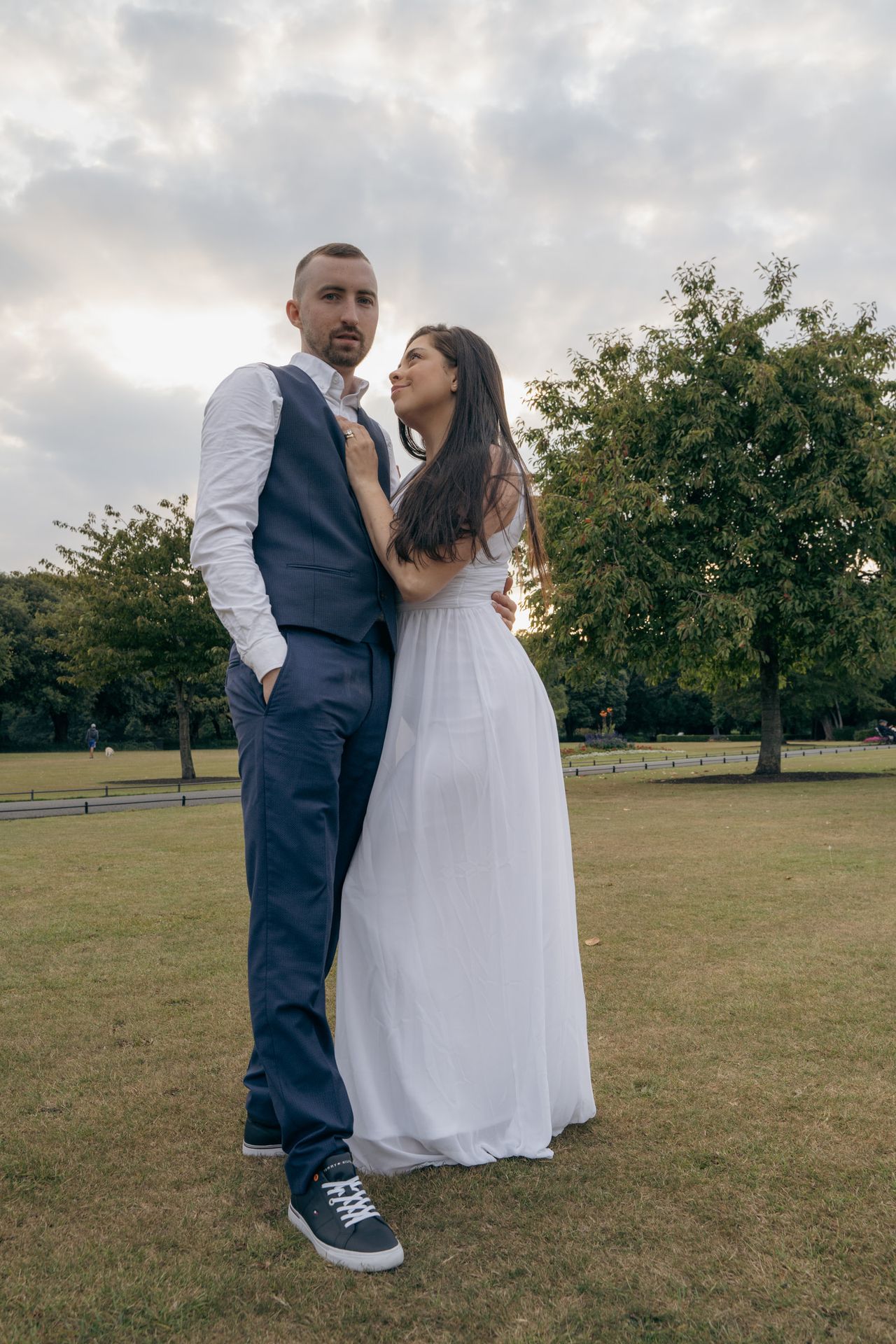 From below on an open lawn under a cloudy sky, now in a long white dress and a navy waistcoat.