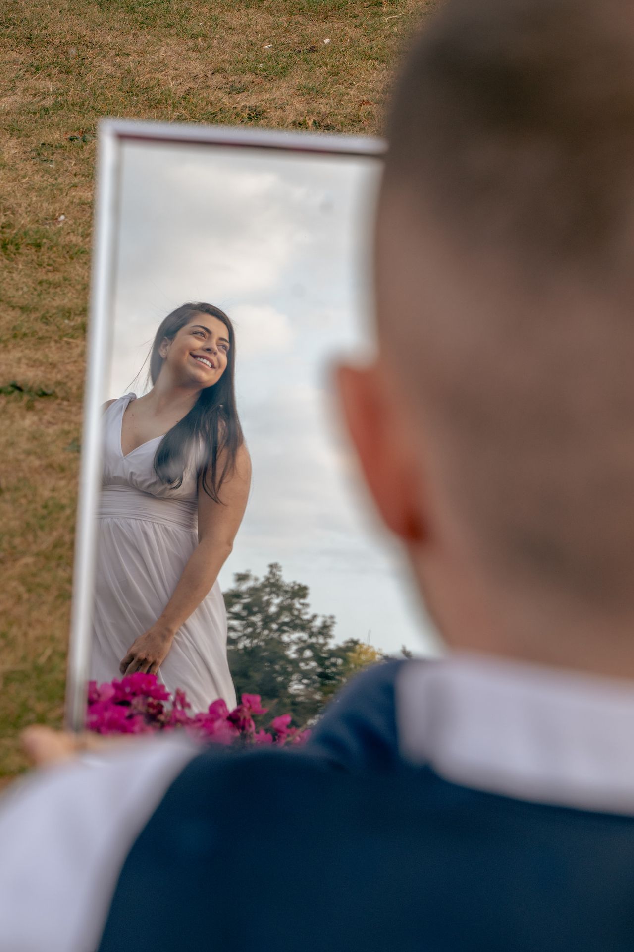 A mirror held up over the grass reflects the long white dress and a smile; the one holding it is seen from behind.