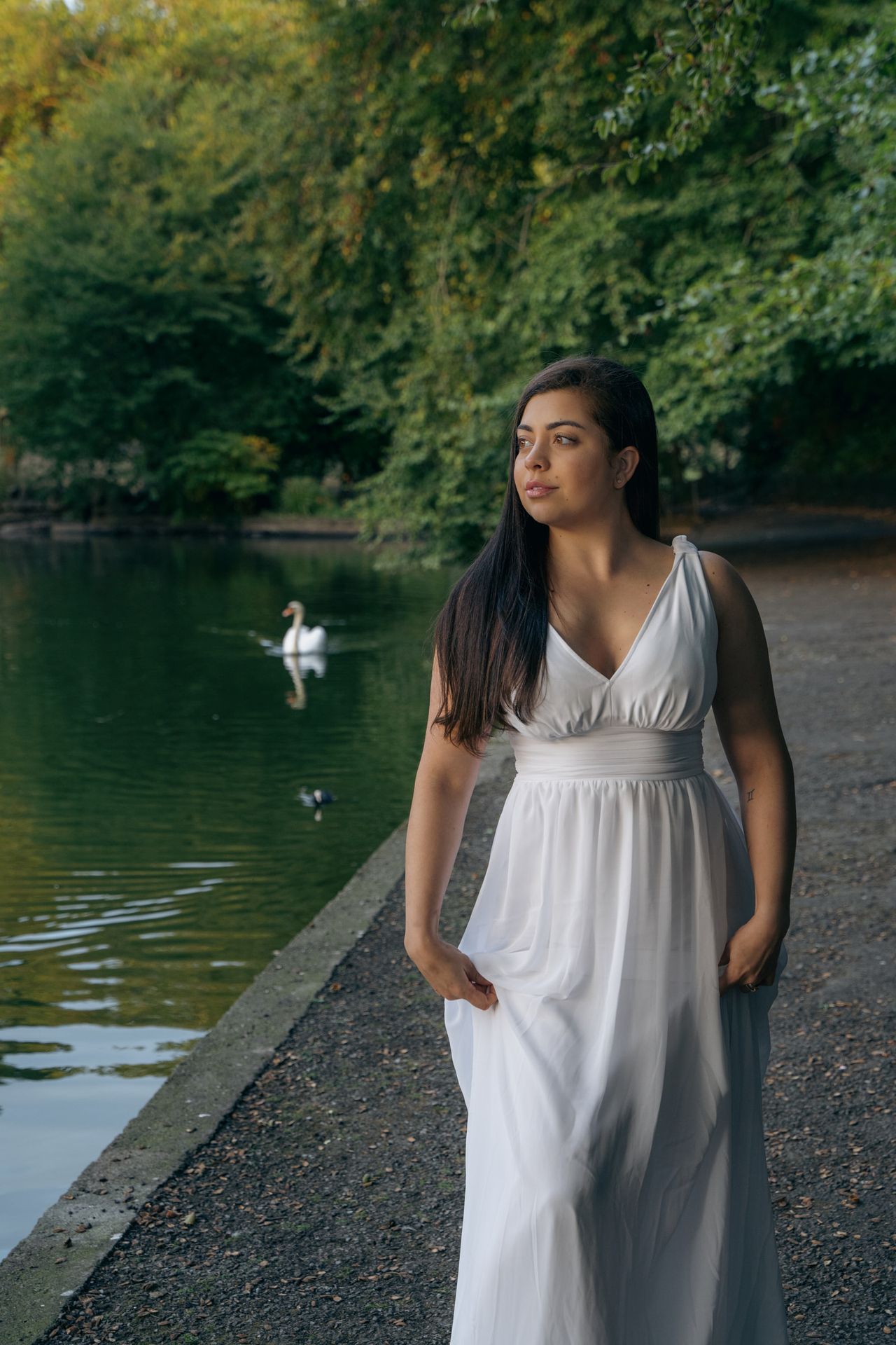 Walking along a pond path in a long white dress, a swan on the water behind.