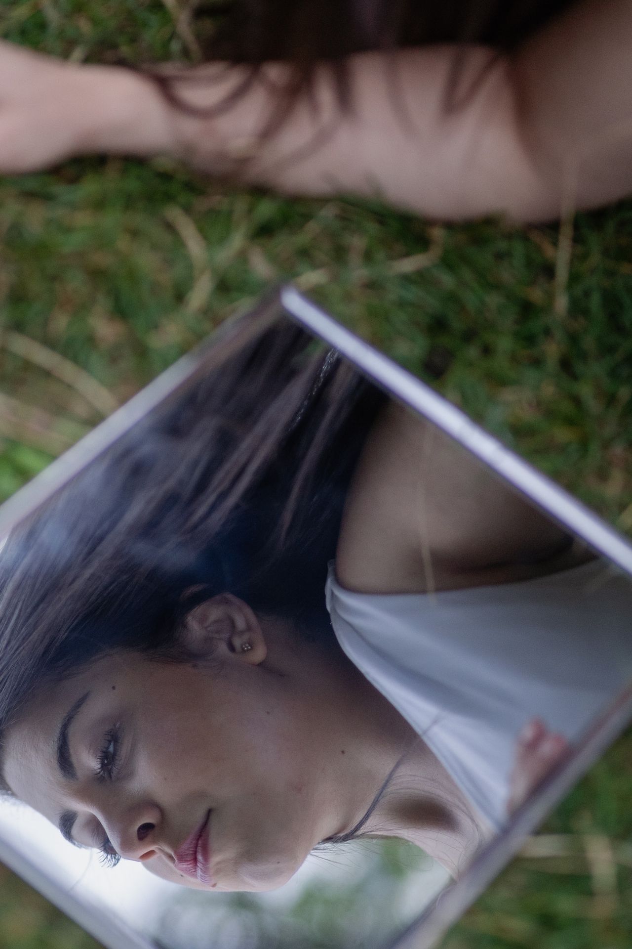 A mirror lying in the grass reflects a face looking down, framed by long dark hair.