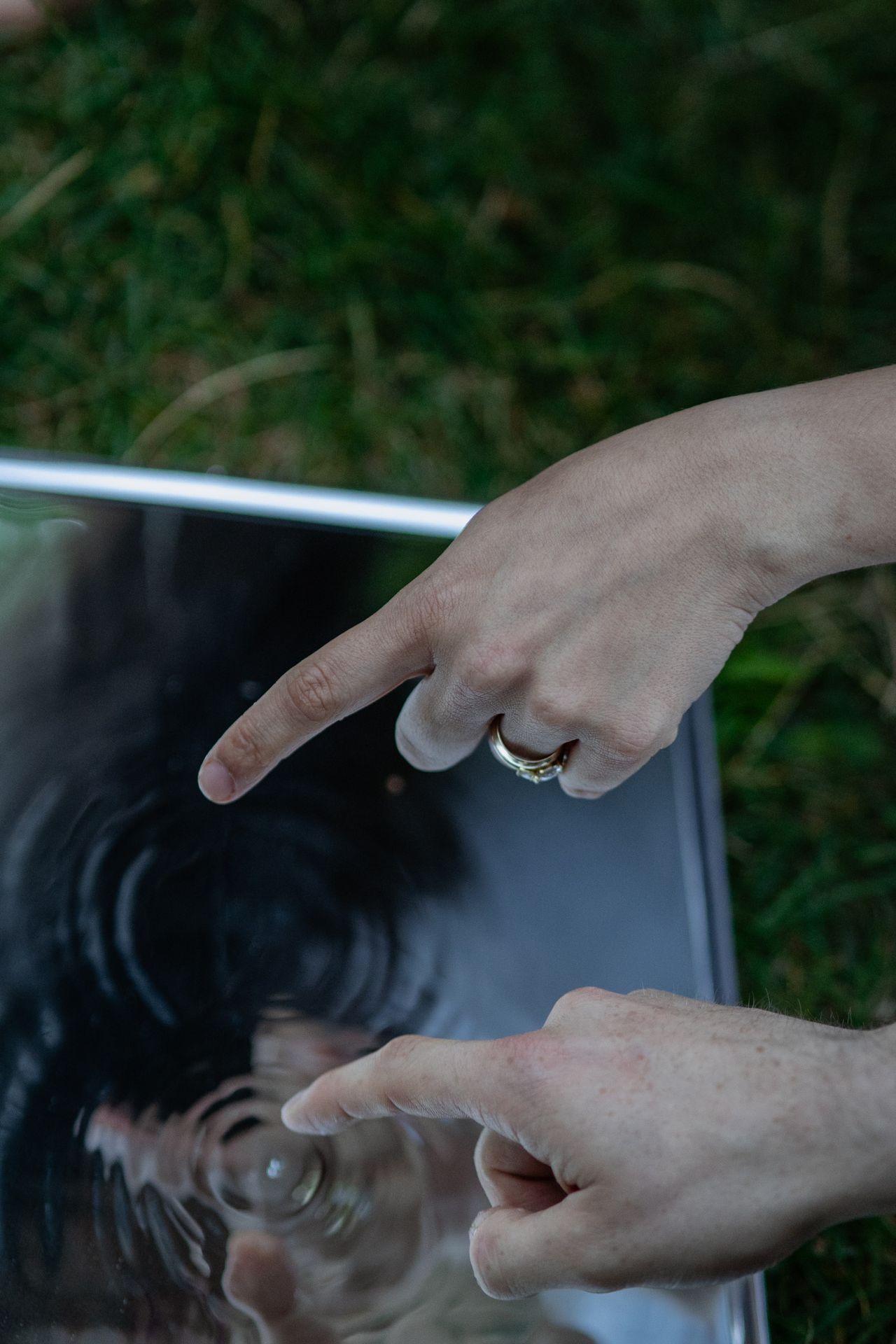 Two fingers point at the mirror in the grass, a ring on one of the hands.