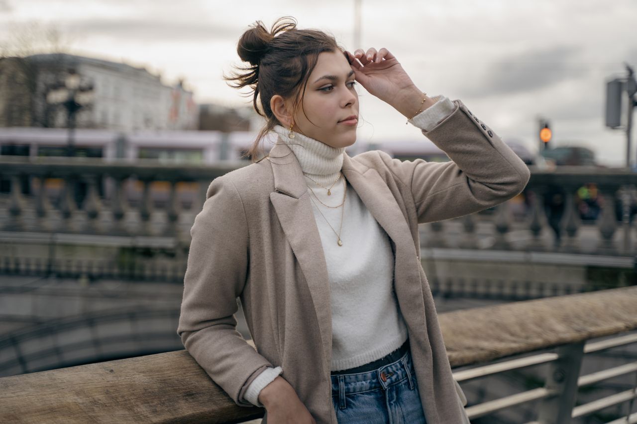 Leaning on a bridge rail in a camel coat, one hand in the hair, a tram crossing behind.