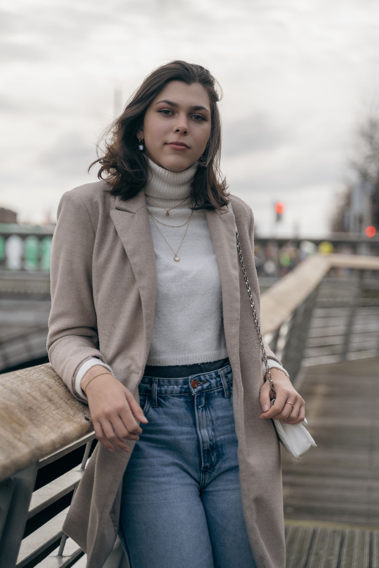 Leaning back against a footbridge rail in a white polo neck and jeans, under a cloudy sky.