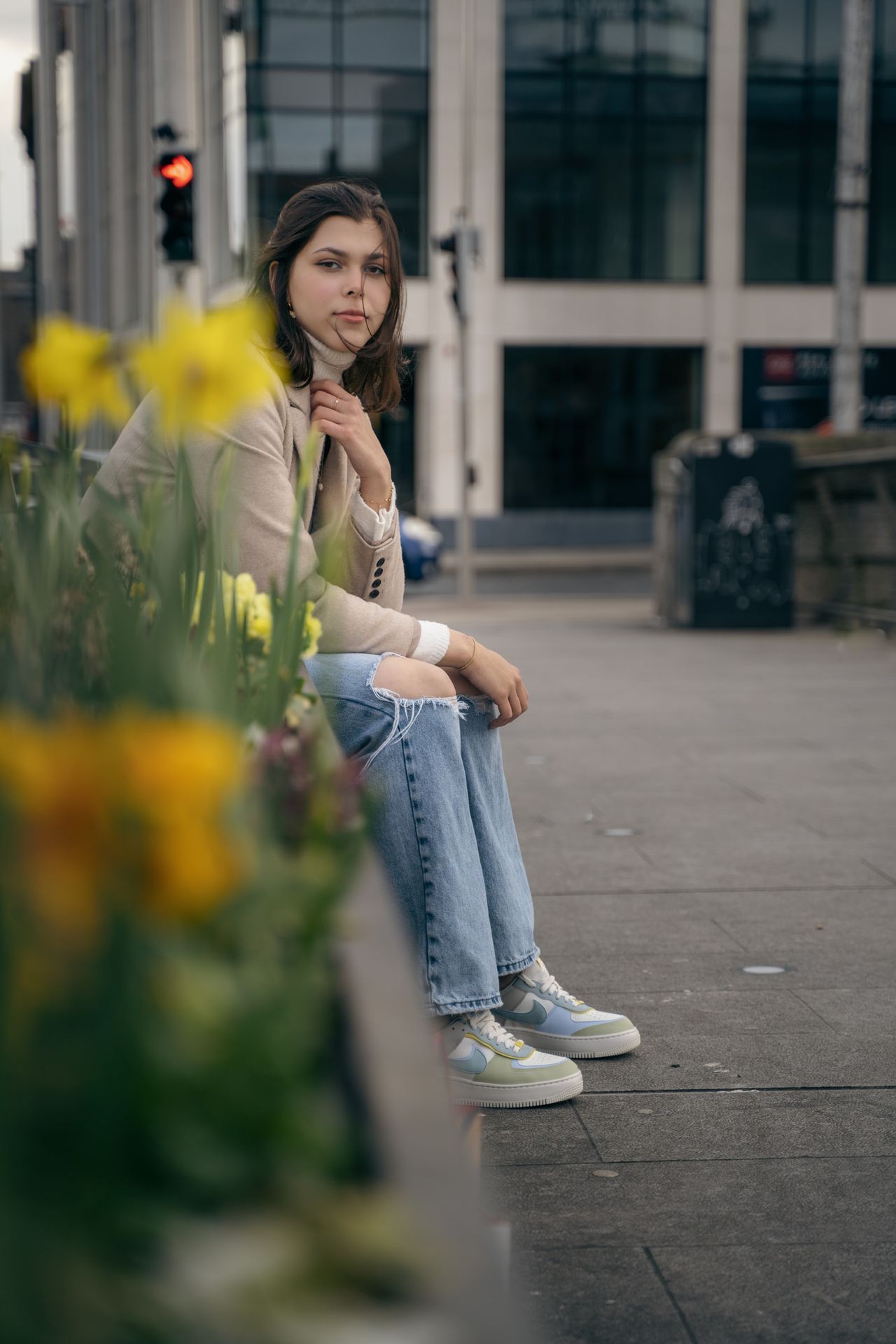 Sitting on a low wall behind yellow daffodils, a glass office building behind.