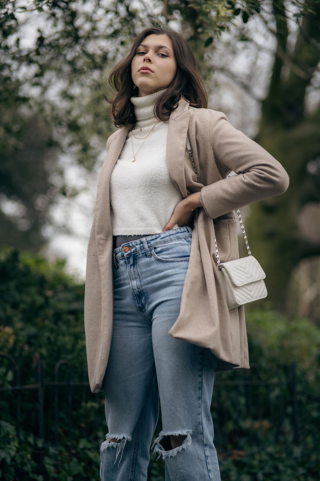Hand on hip in front of dark green trees, the coat open over a white jumper.