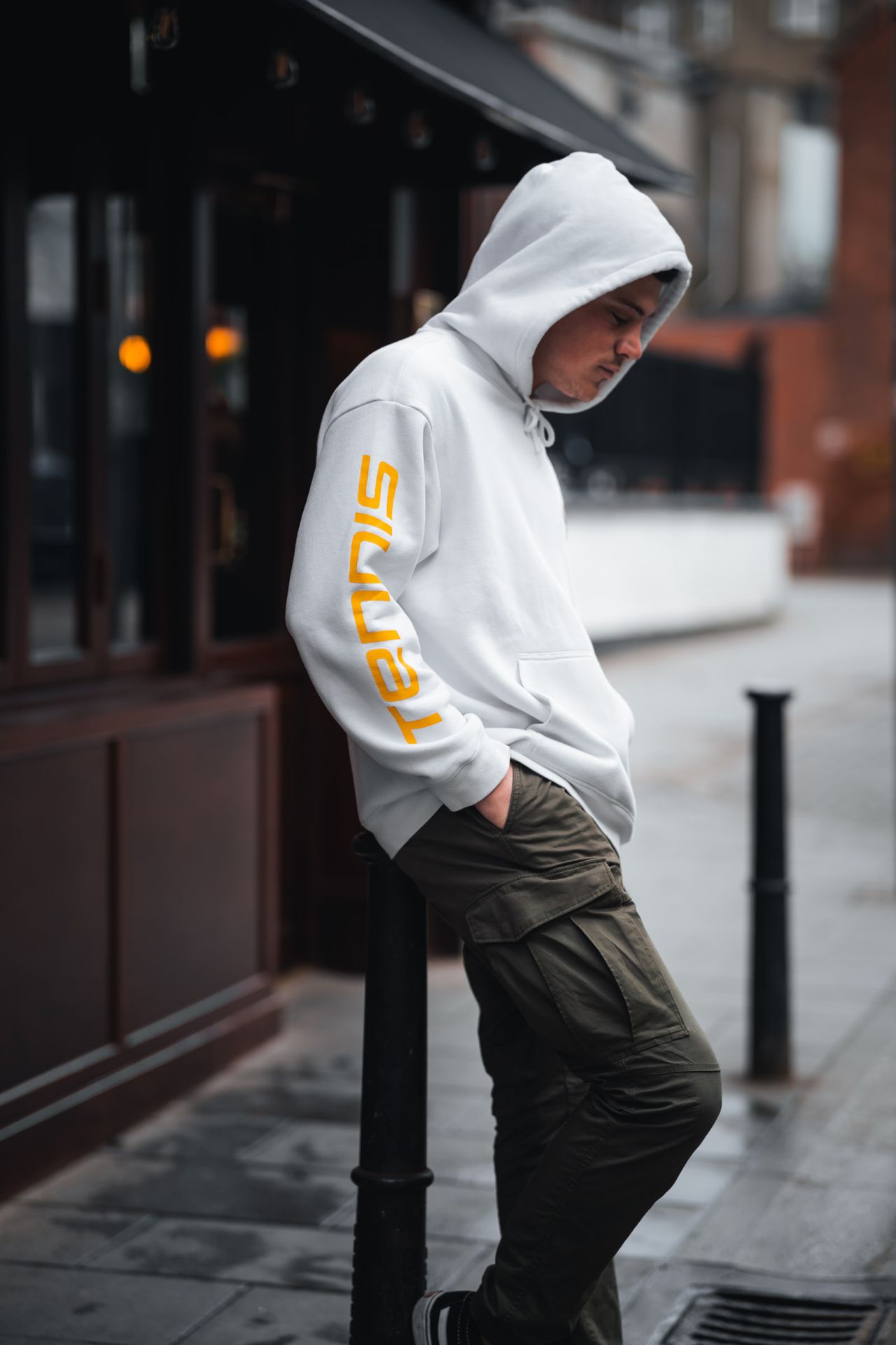 Hood up, leaning on a bollard outside a dark shop front, eyes down.