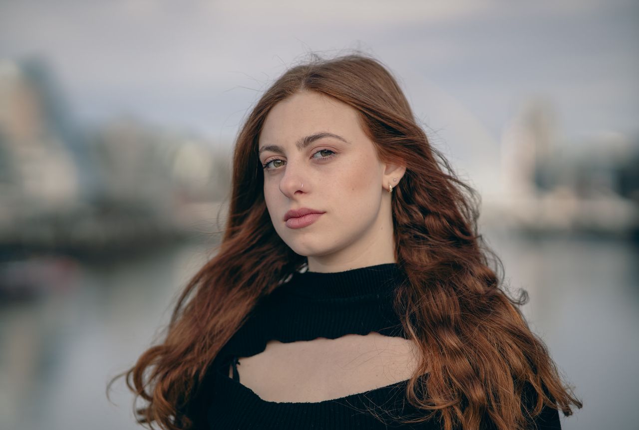 Close portrait by the river, long red hair over a black top, the city soft behind.