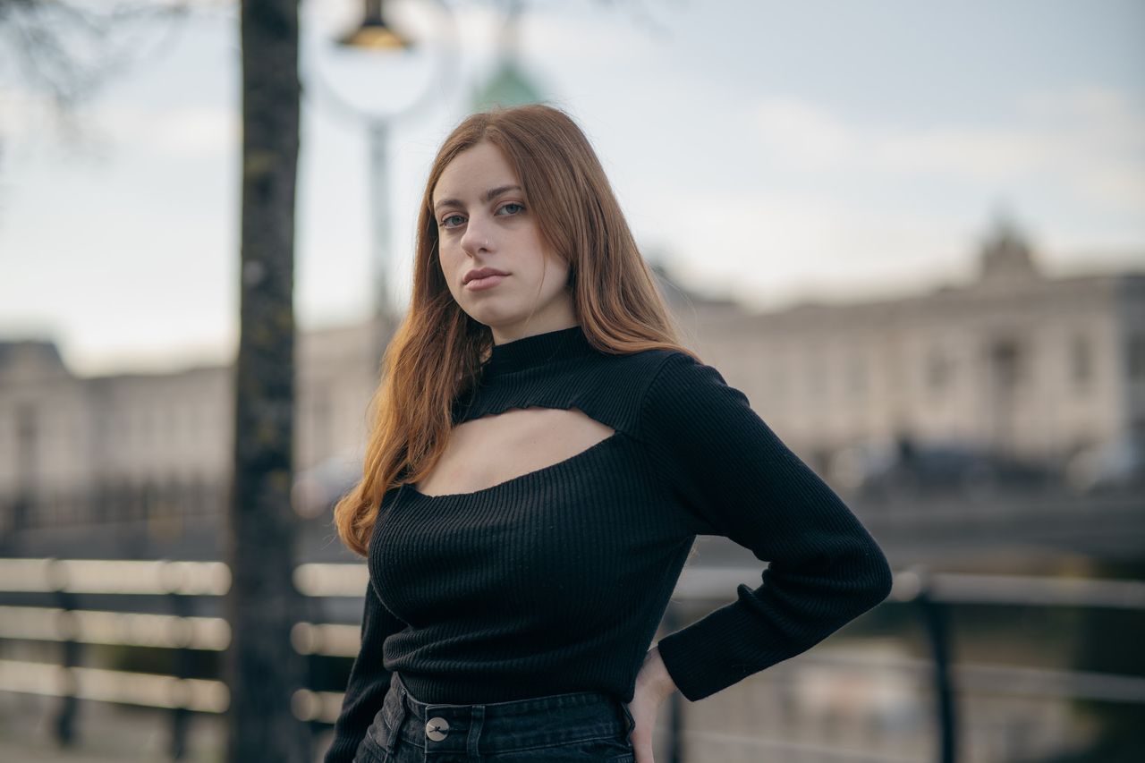 Hand on hip by the river railings, a domed building blurred across the water.