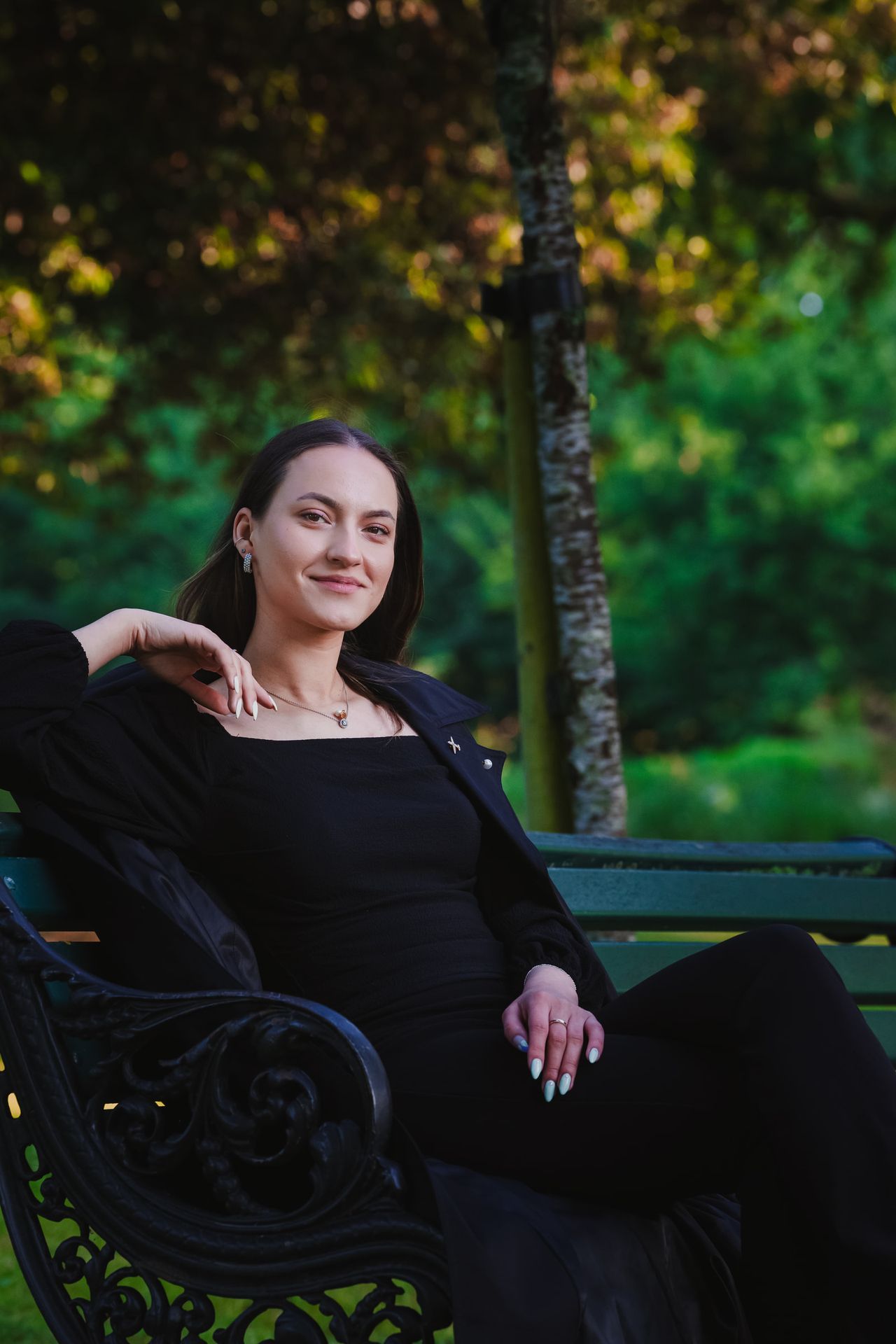 Sitting back on an iron park bench in a long black coat, smiling at the camera.