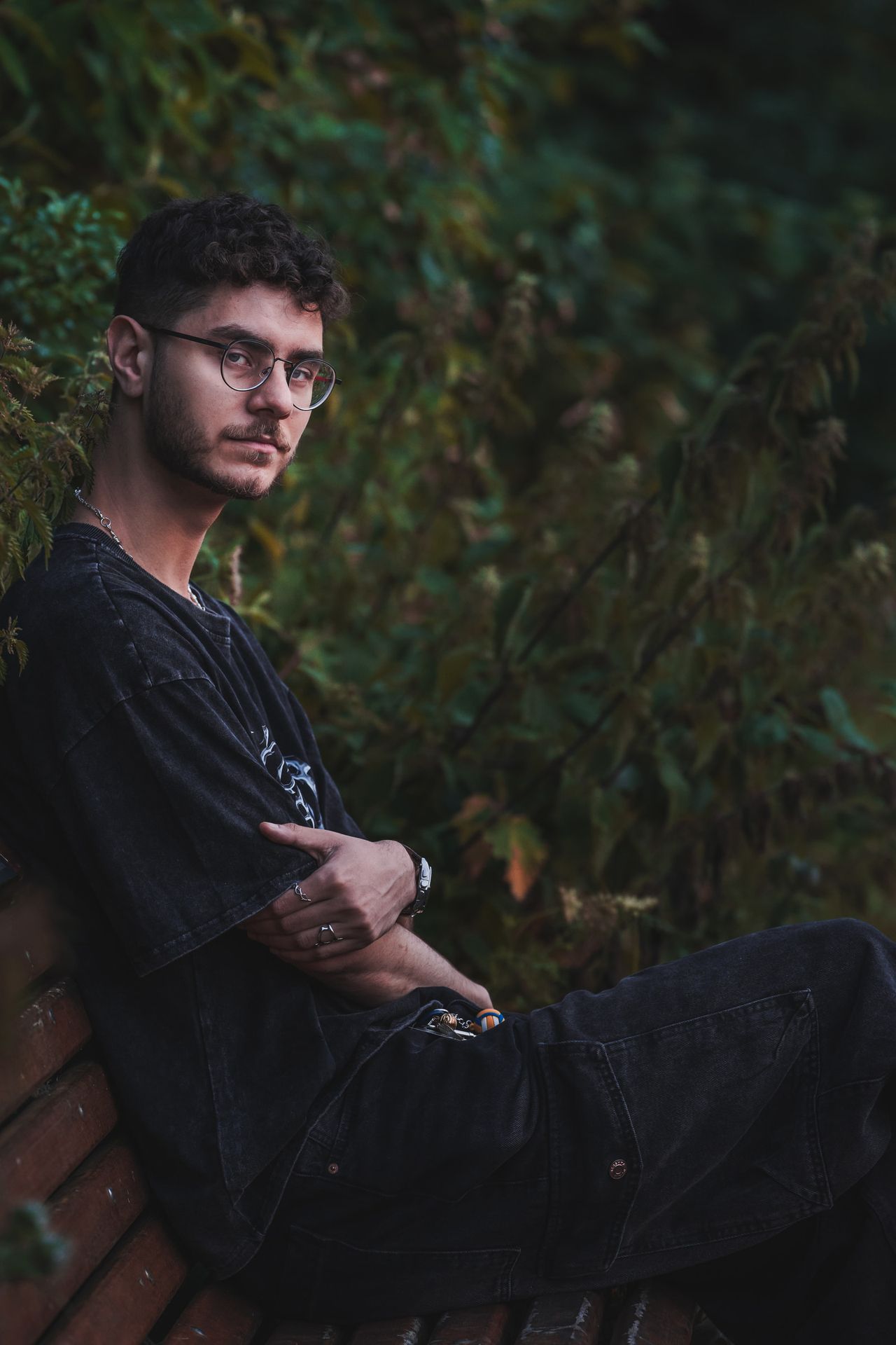 Sitting on a bench among dark leaves, arms crossed, wearing glasses.