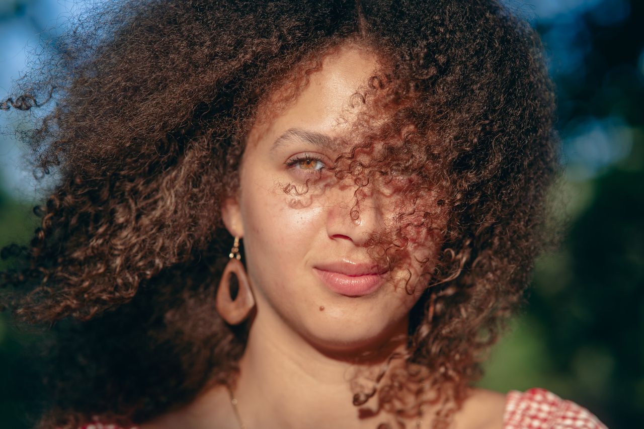 Close-up with curls blowing across the face, a wooden earring catching the light.