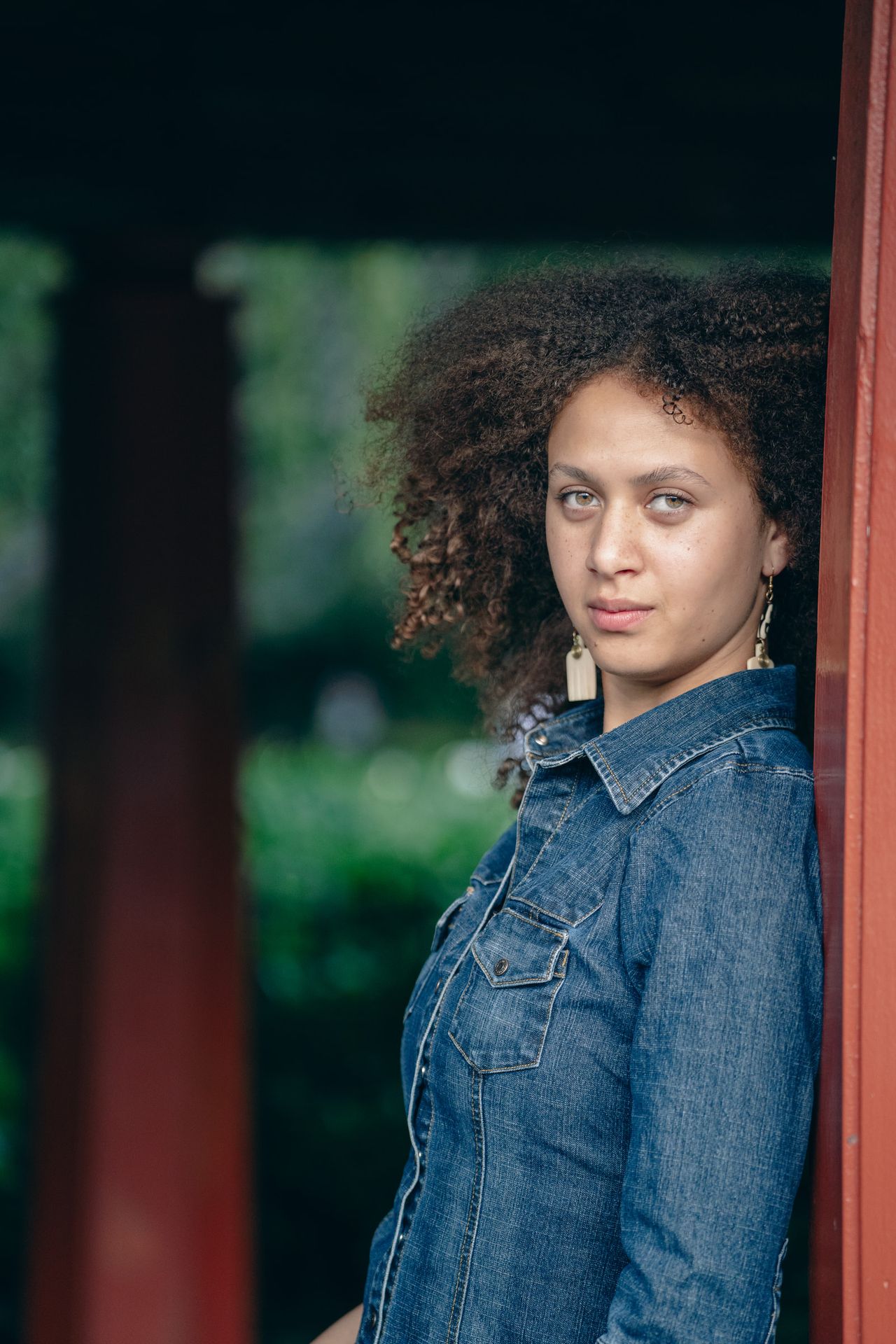 In a denim shirt, leaning on a red pillar and looking straight at the camera.