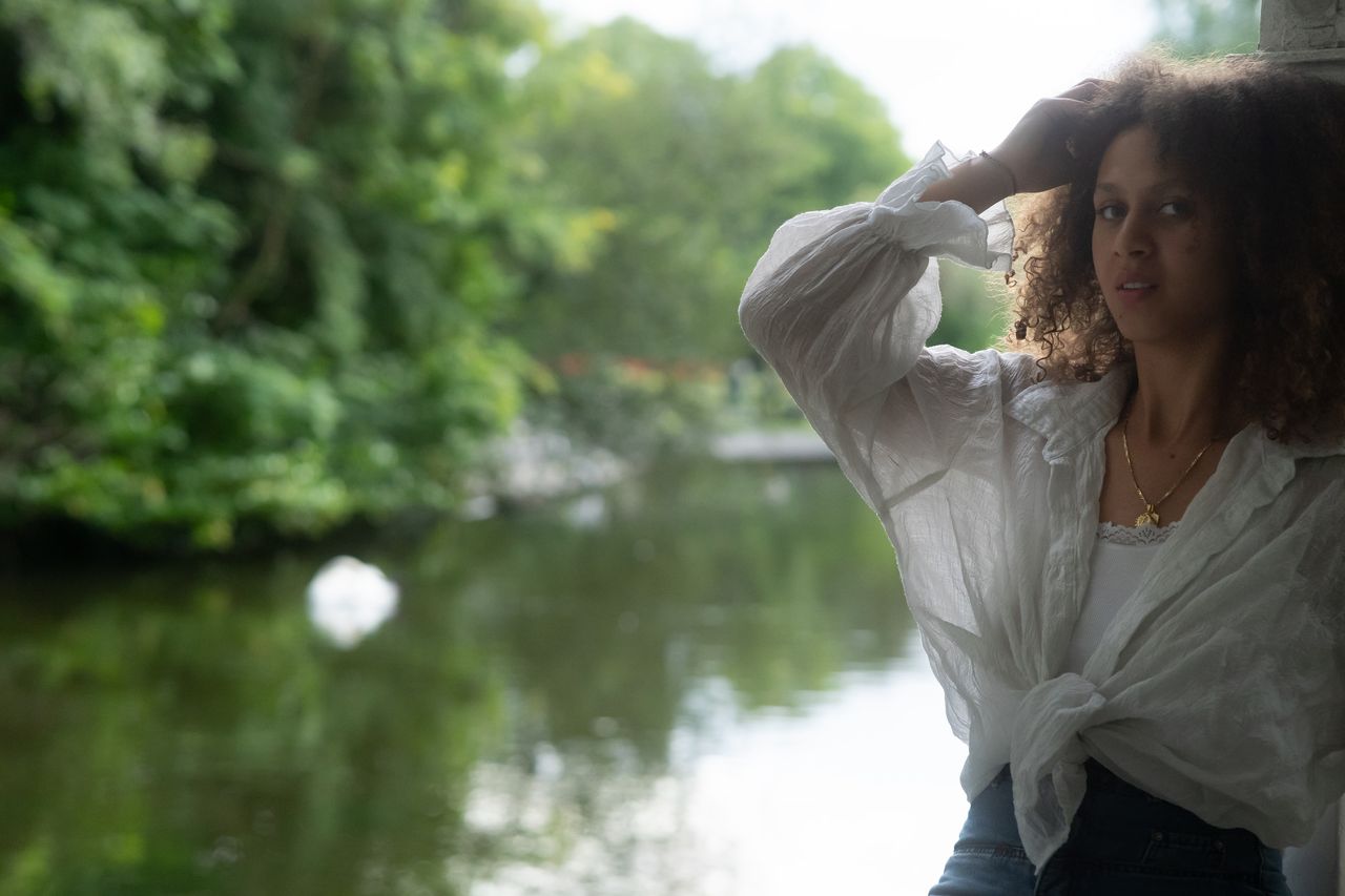 One hand in a head of curls, looking at the camera beside a pond framed by summer trees.