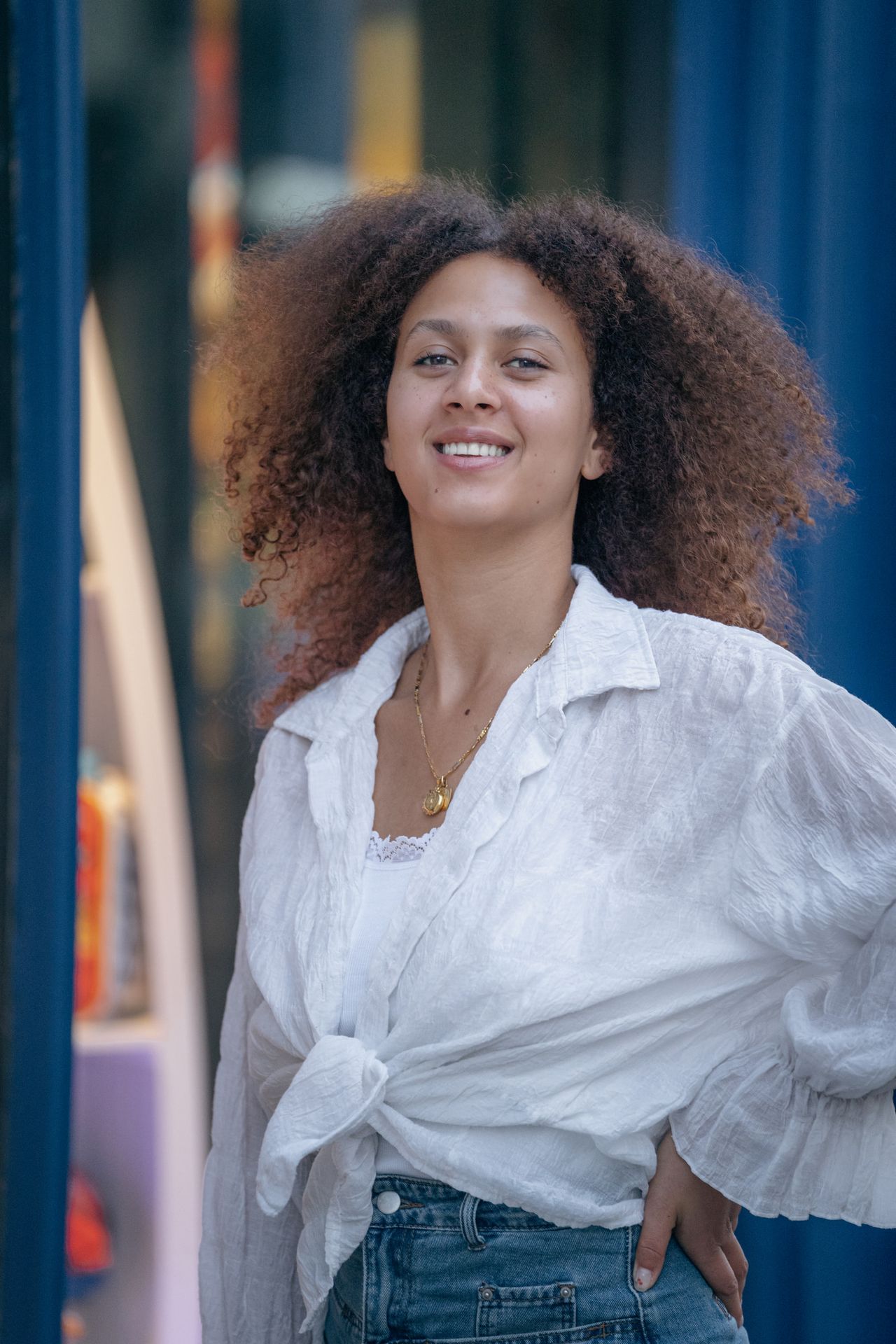 Smiling in a white shirt in front of a blue door, one hand on the hip.