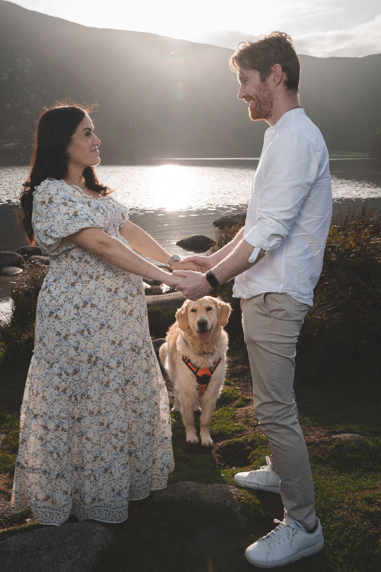 The couple hold hands on the shore of a mountain lake, a golden-haired dog sitting between them.