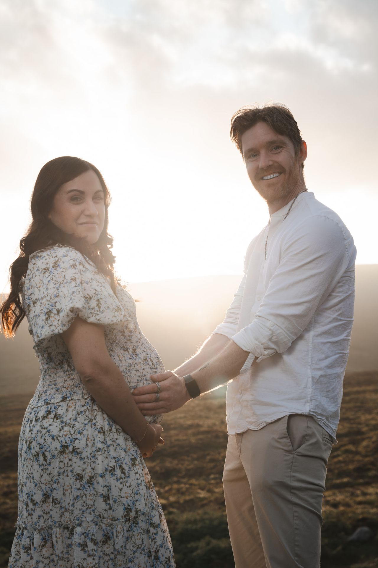 Side by side on the hillside against the setting sun, one hand on the bump, both smiling at the camera.