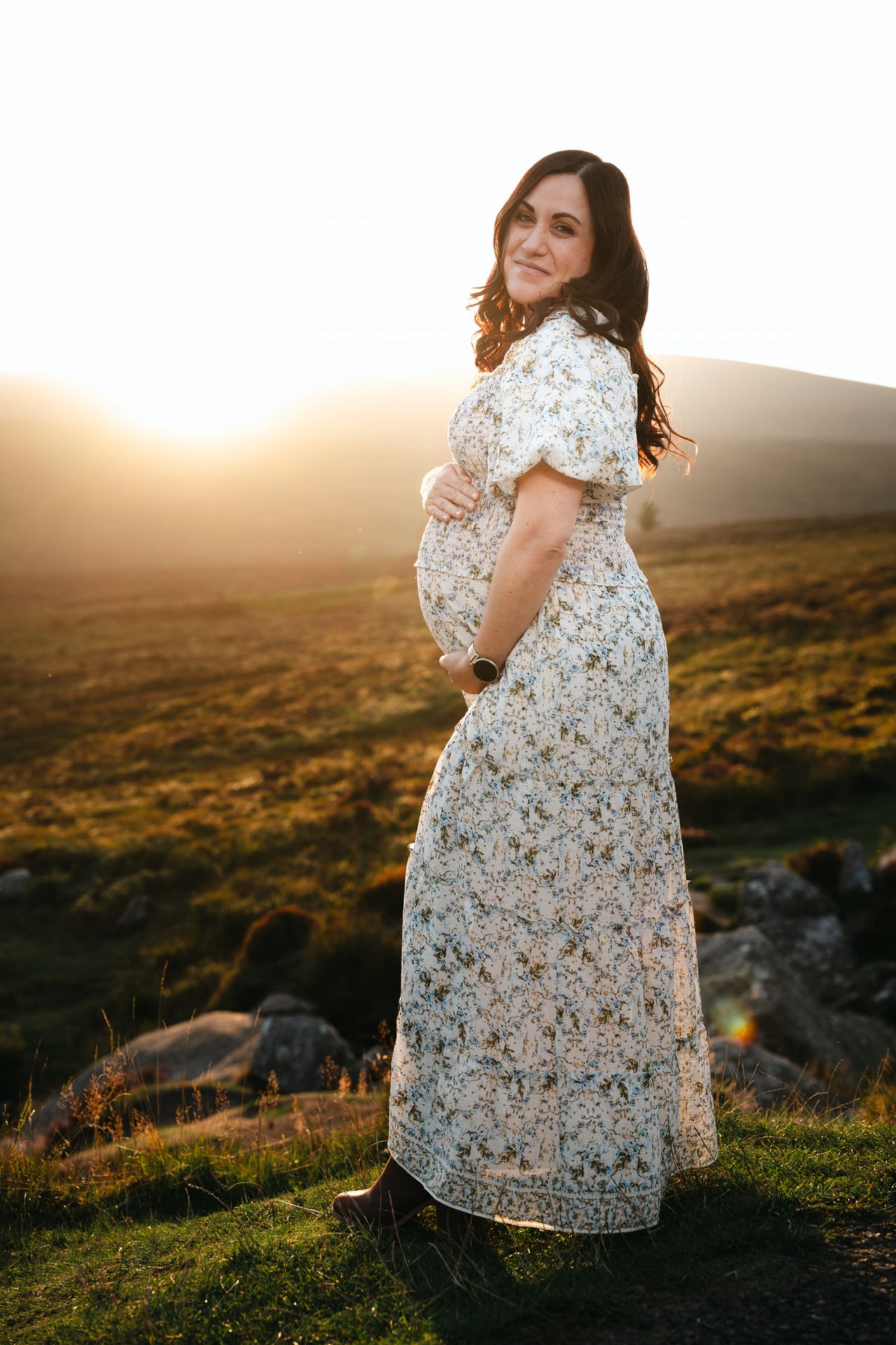Standing on the hillside with the sun low behind, both hands around the bump, smiling over one shoulder.