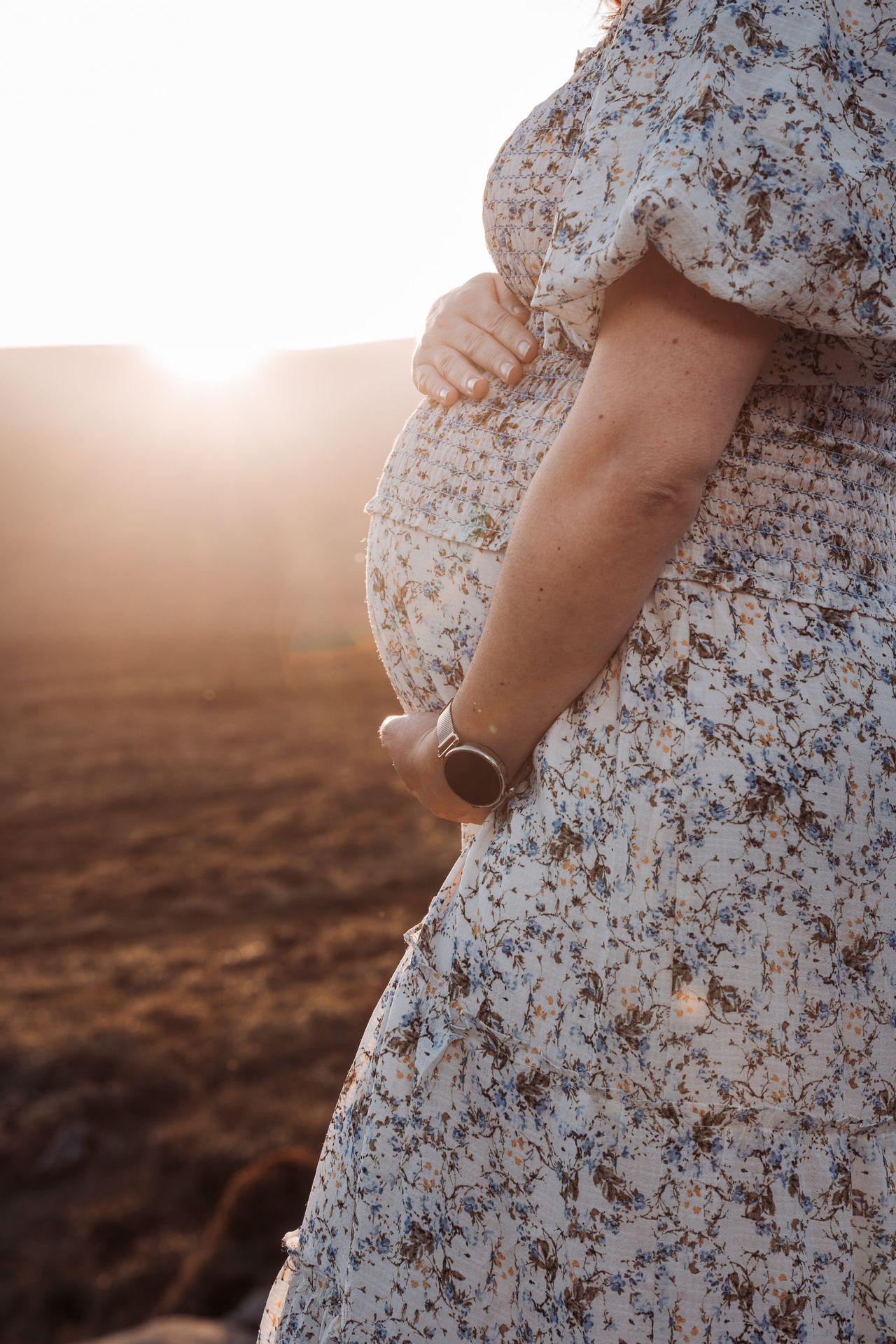 Close-up of the bump under a floral dress, one hand resting on it as the sun sets.